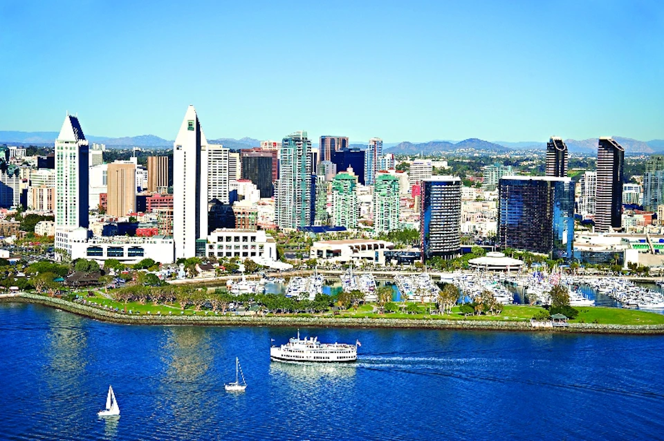 City skyline with various high-rise buildings, a waterfront park, and boats on a blue river under a clear sky.