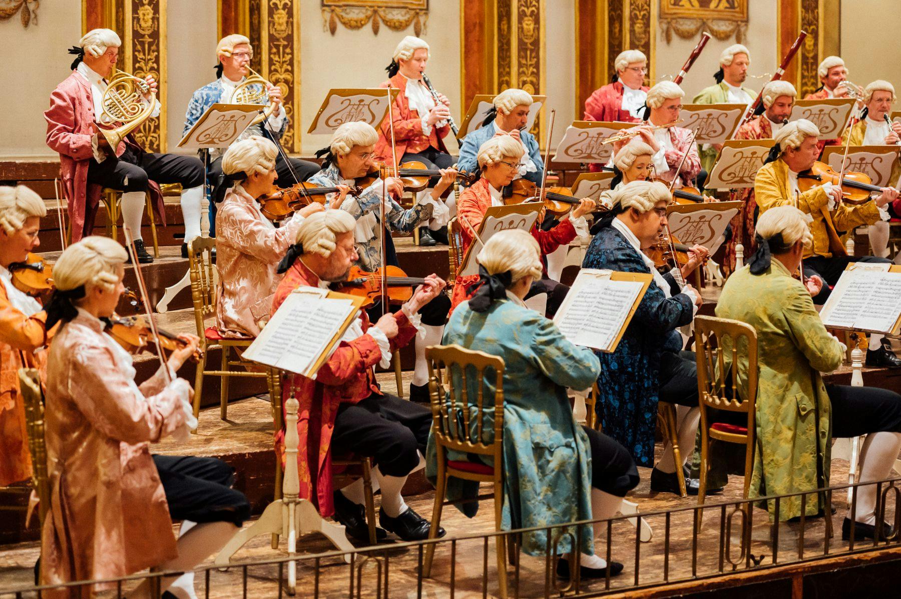 Orchestra dressed in period costumes with sheet music on stands performs on stage in an ornate hall.