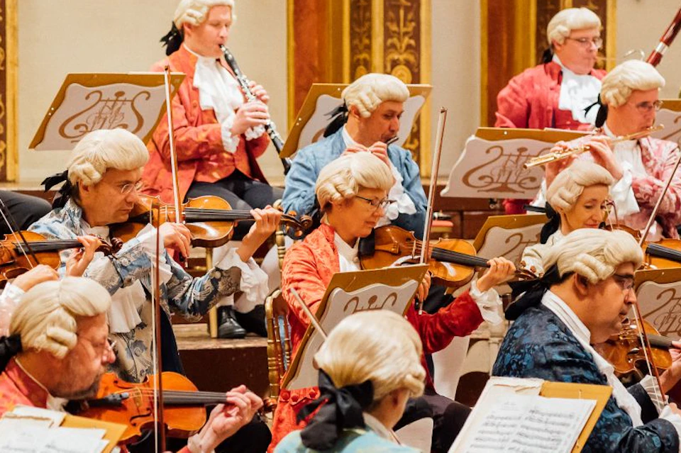 Orchestra dressed in period costumes with sheet music on stands performs on stage in an ornate hall.