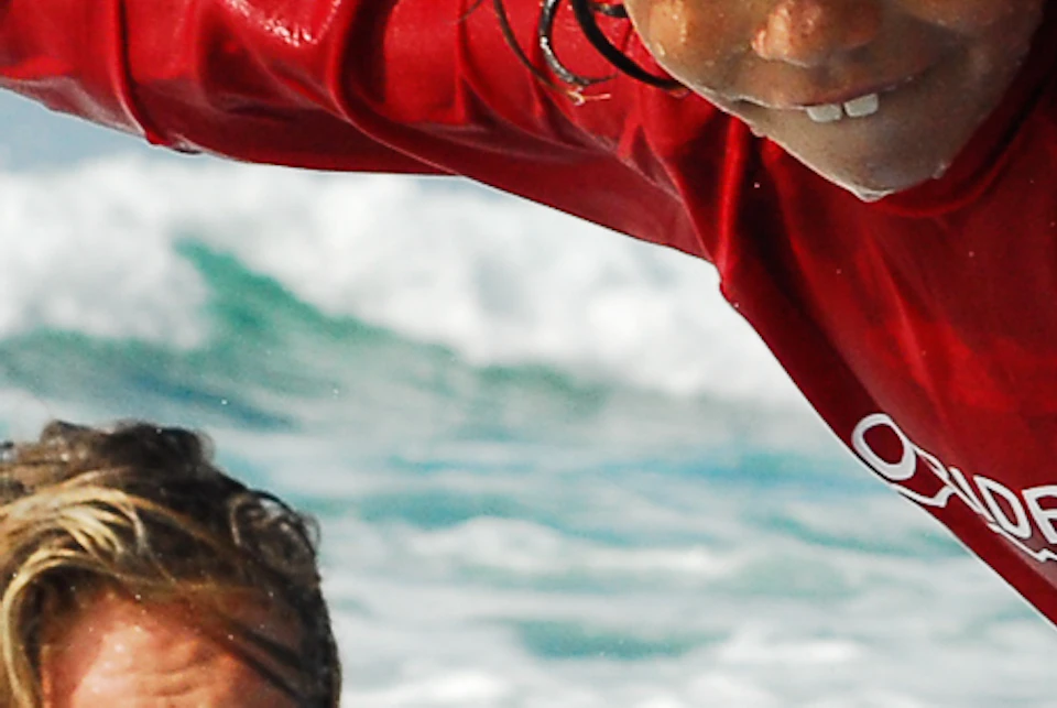 A child in a red wetsuit balances on a surfboard while an adult in the background watches, both in the ocean waves.