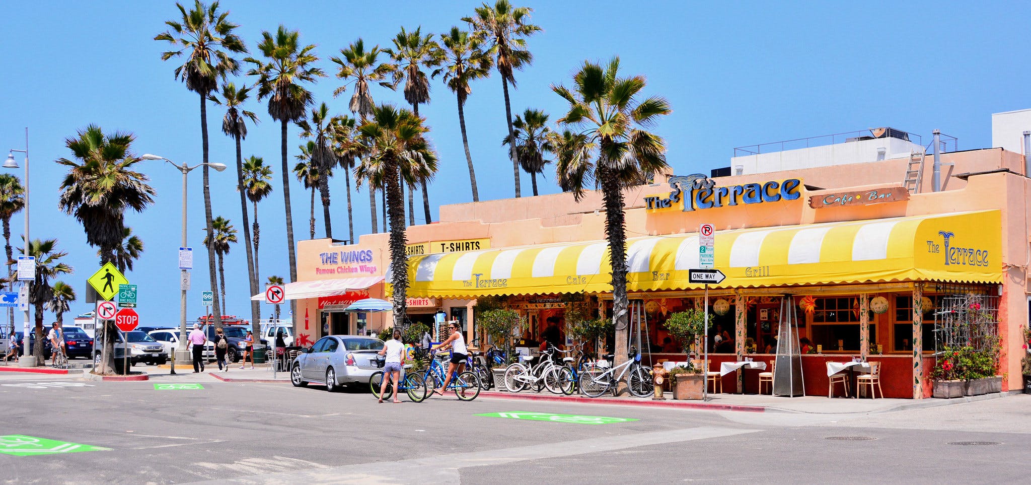 "The Terrace" 카페와 "The Wings" 매장을 포함한 상점들이 있는 야자수가 늘어선 거리. 걷고 자전거 타는 사람들, 주차된 자동차, 맑고 푸른 하늘.