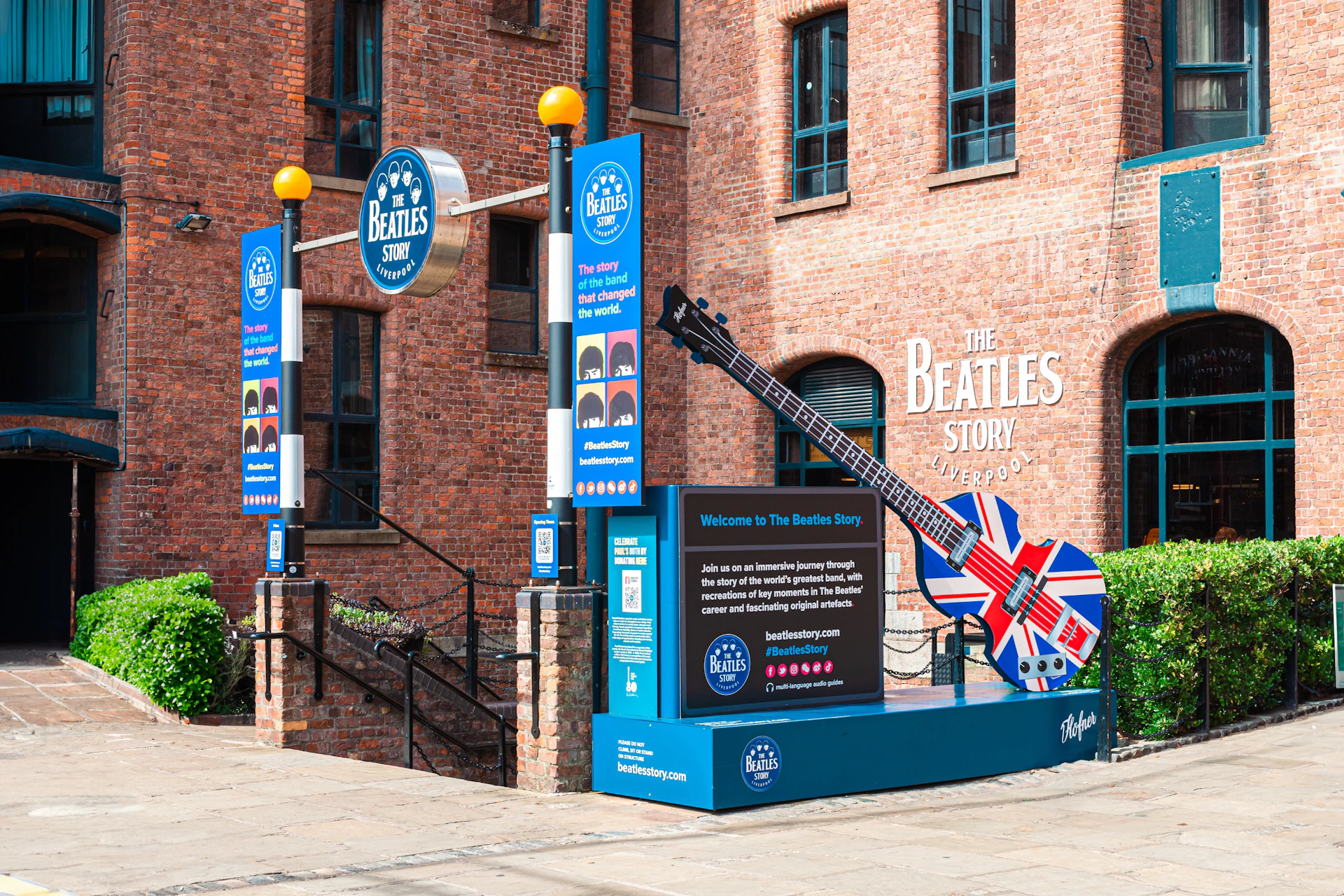 Entrance to The Beatles Story museum in Liverpool, featuring posters, signs, and a guitar sculpture with a Union Jack design.