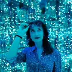 A woman in a sparkly shirt stands against a backdrop of blue, shimmering lights, adjusting her sunglasses while looking upward.