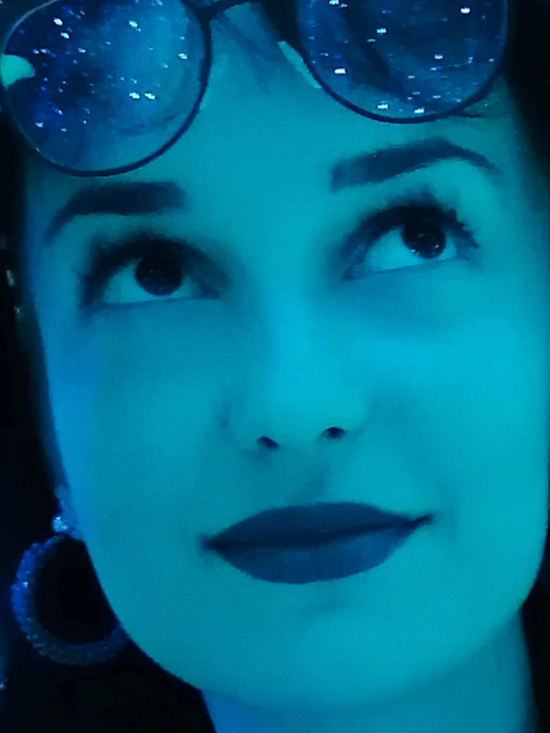 A woman in a sparkly shirt stands against a backdrop of blue, shimmering lights, adjusting her sunglasses while looking upward.