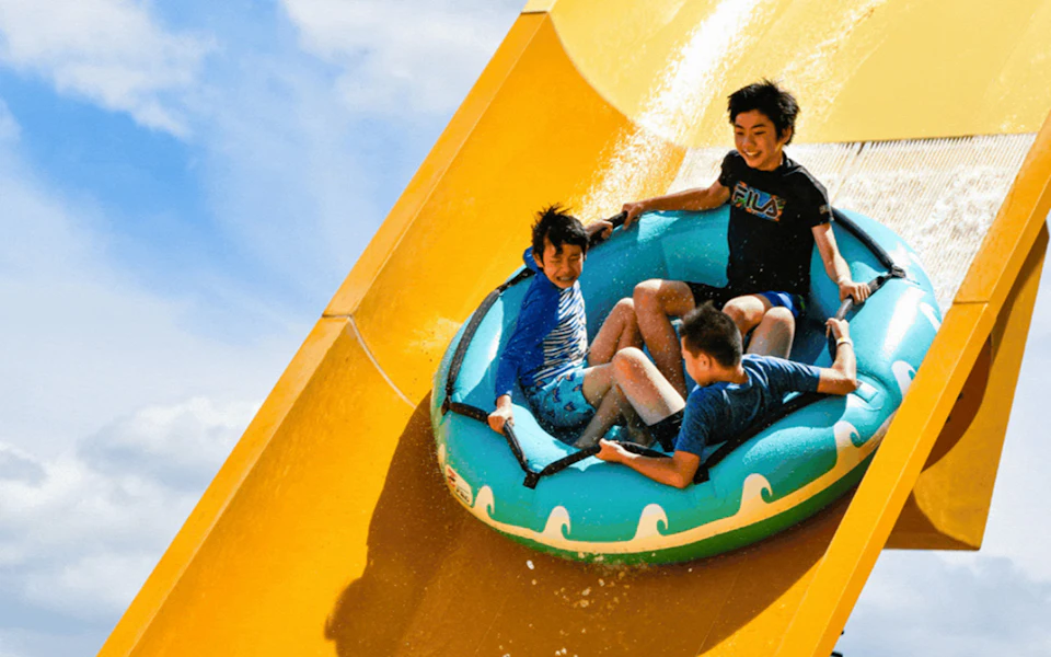 Three individuals riding a blue inflatable raft down a yellow water slide on a sunny day.