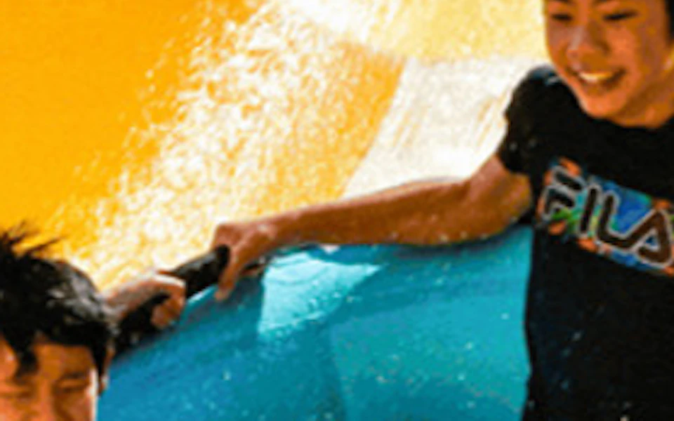 Three individuals riding a blue inflatable raft down a yellow water slide on a sunny day.