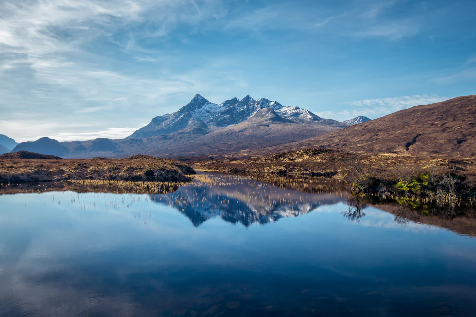 Snow-capped mountains reflect in a calm lake under a clear blue sky, surrounded by dry, rocky terrain.