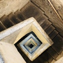 An overhead view of a square spiral staircase made of worn concrete. The stairs descend in a geometric pattern.