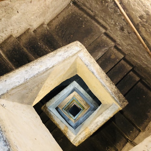 An overhead view of a square spiral staircase made of worn concrete. The stairs descend in a geometric pattern.