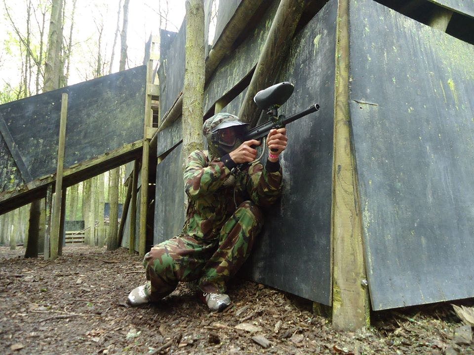 Ένα άτομο με εξοπλισμό παραλλαγής σκύβει πίσω από ένα ξύλινο φράγμα σε μια δασώδη περιοχή, κρατώντας ένα όπλο paintball.