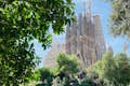 Vue de la Sagrada Familia depuis un parc voisin, encadrée par une verdure luxuriante et mettant en valeur ses flèches imposantes.