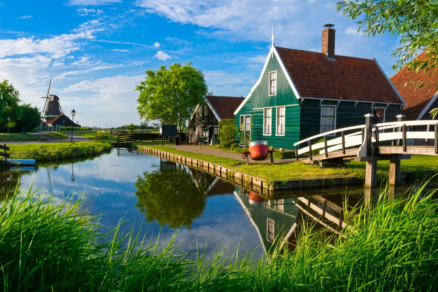 穏やかな運河沿いにある赤い屋根のグリーンハウス、白い歩道橋、青い空と散在する雲の下にある緑豊かな緑。