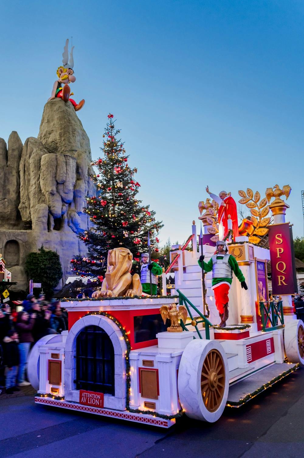 A festive parade floats with Roman-themed decorations, a Christmas tree, and a large rock formation in the background.