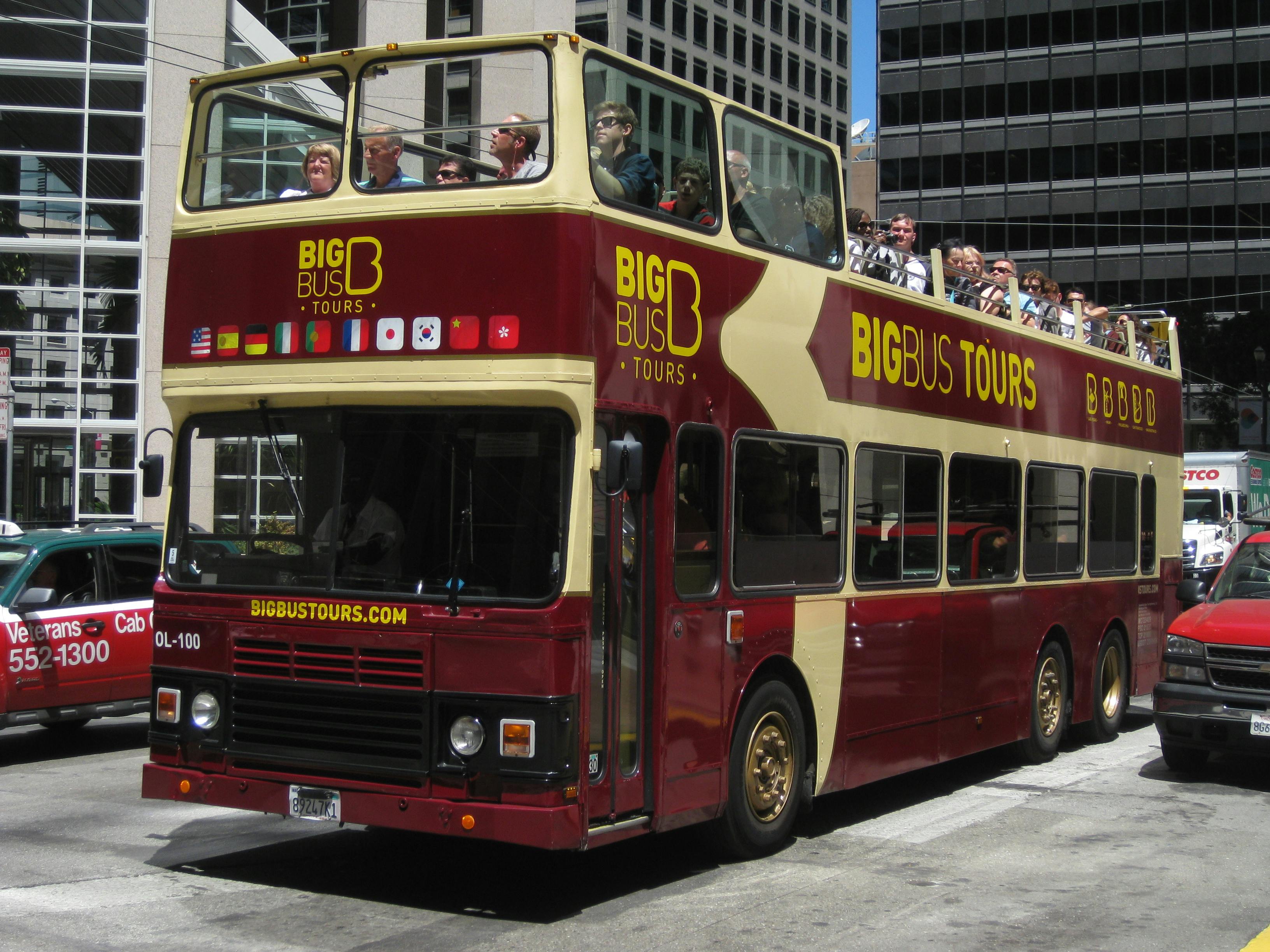 Ein rot-gelber doppelstöckiger Tour-Bus mit Fahrgästen auf dem offenen Oberdeck, geparkt auf einer Stadtstraße.
