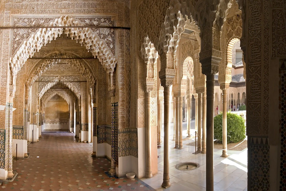 A richly decorated corridor with intricate arches and columns, featuring Islamic-style geometric designs and a tiled floor.