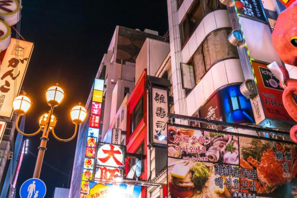 City street at night with brightly lit signs in various languages and a streetlamp in the foreground.