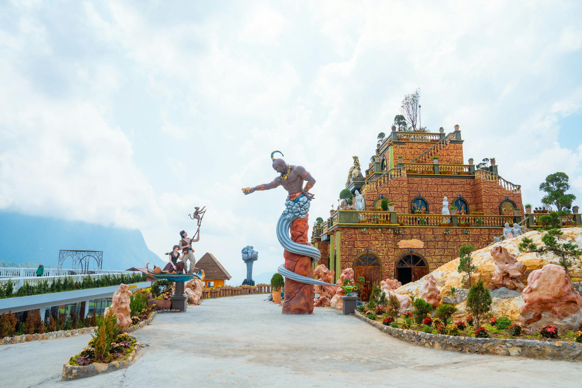 A colorful fantasy-themed outdoor park with statues, a decorated building, and landscaped pathways under a cloudy sky.