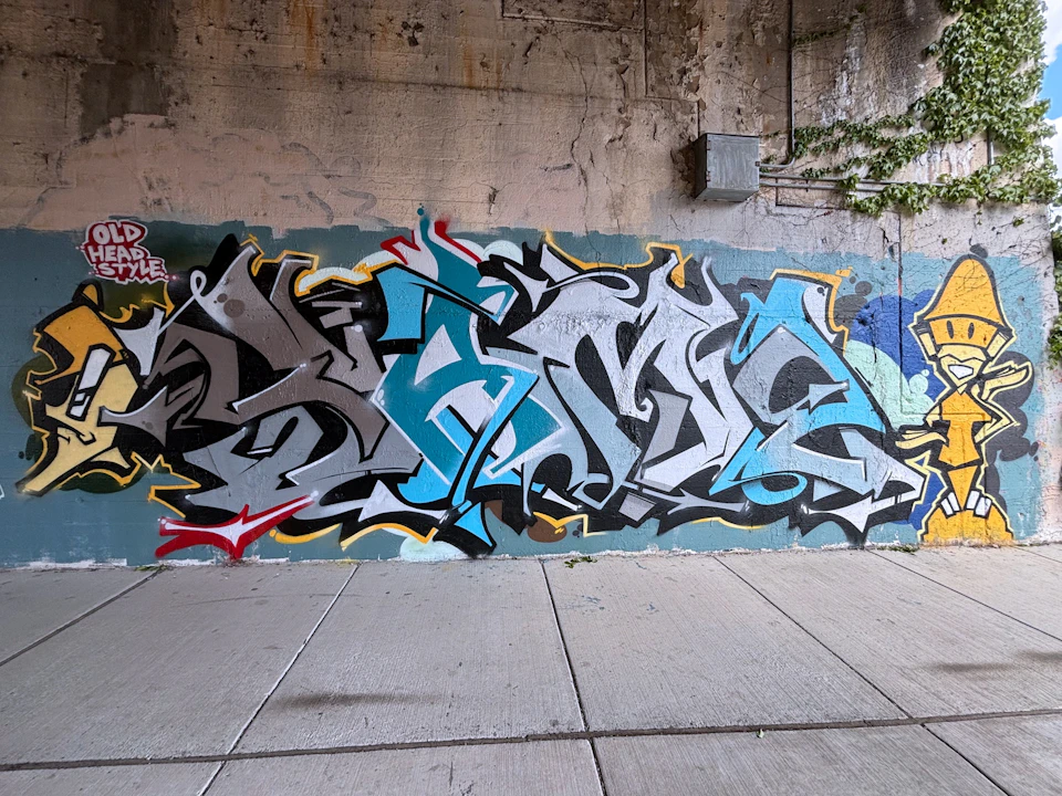 A colorful graffiti mural with abstract shapes and letters on a concrete wall under an overpass.