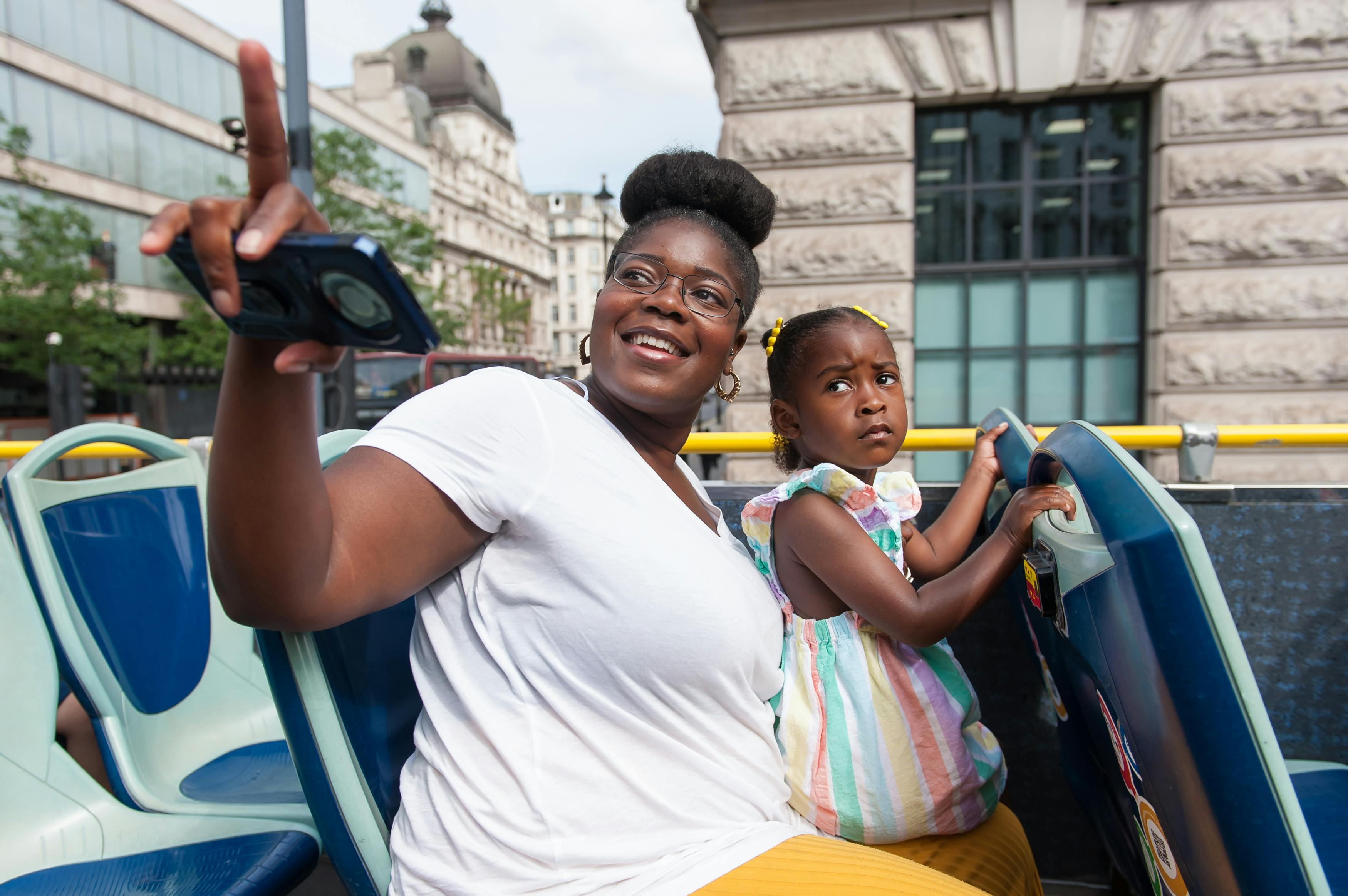 한 여성과 어린 소녀가 2층 버스 꼭대기 데크에 앉아 있습니다. 여자가 웃으며 셀카를 찍고 있다. 소녀는 진지해 보인다.