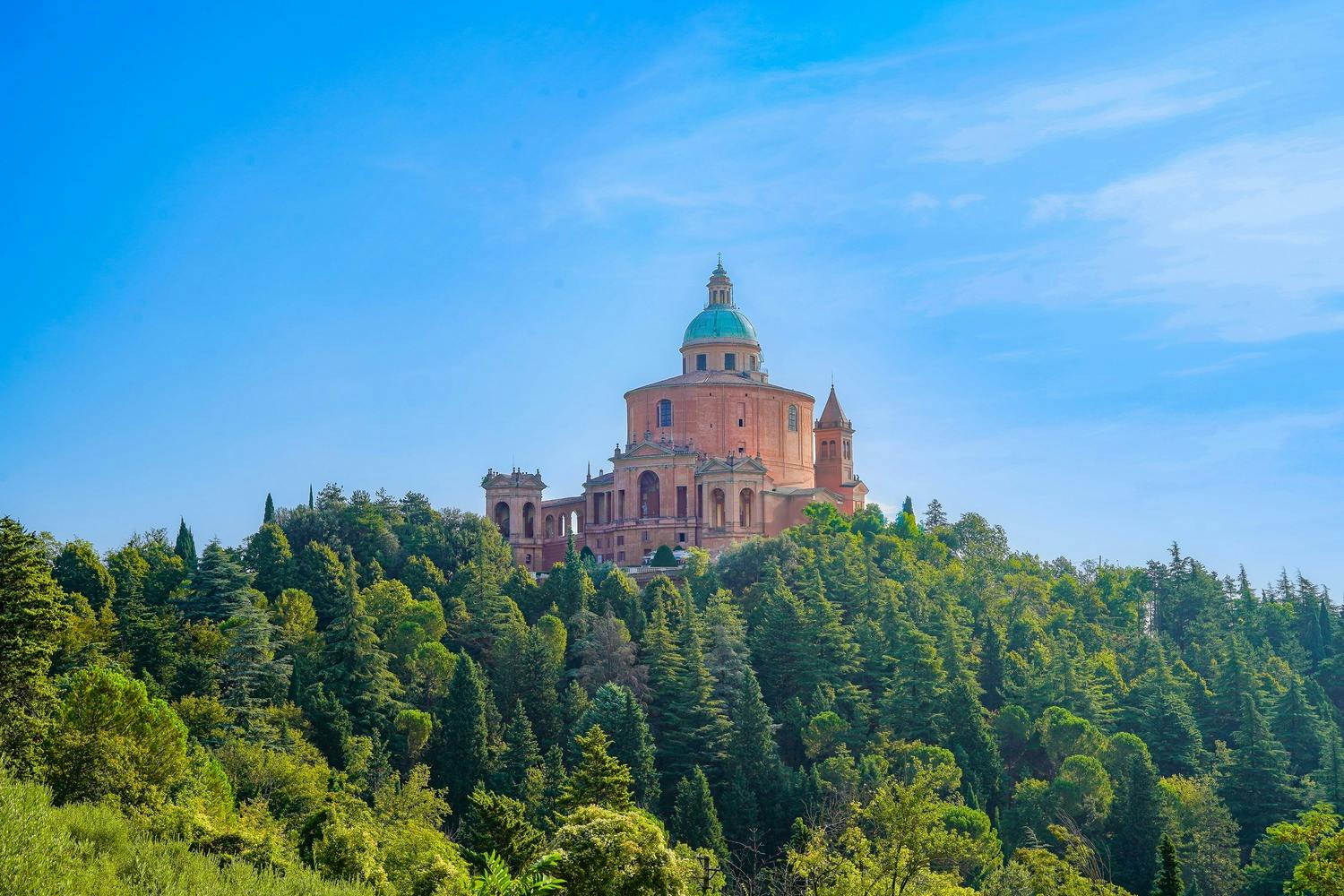 A large, historic building with a central dome and spires surrounded by dense, green trees under a clear blue sky.