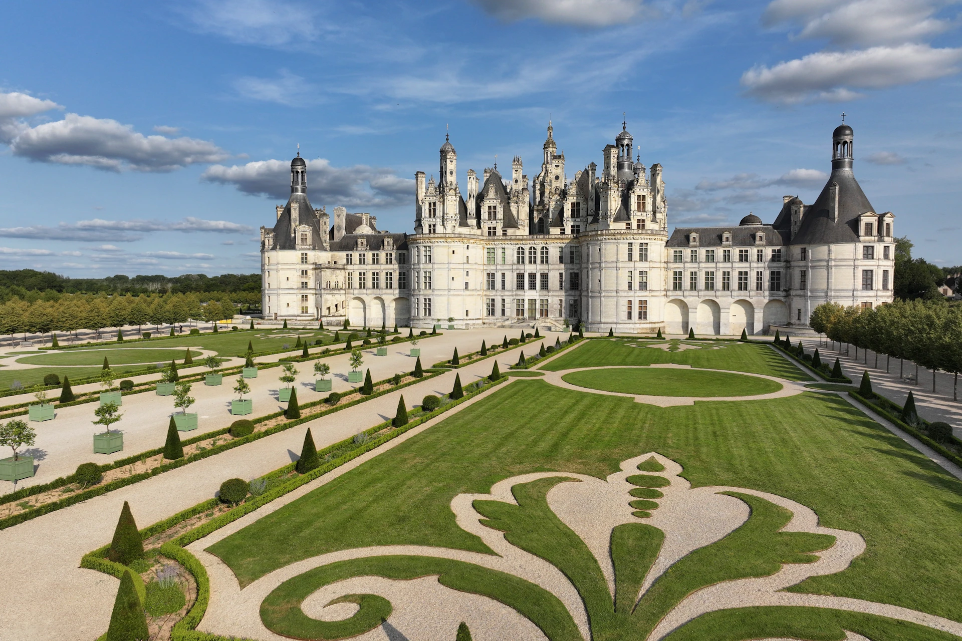 A grand castle with ornate architecture surrounded by manicured gardens under a blue sky with scattered clouds.