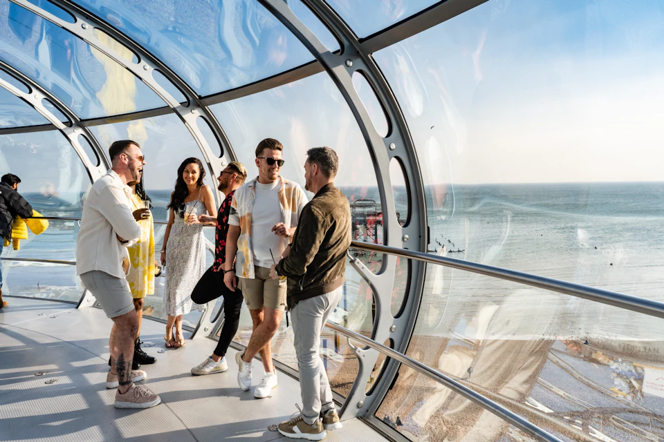 A group of people standing and talking inside a glass-enclosed observation pod with a view of the sea and coastline.