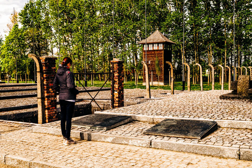 A person stands alone, facing a memorial with plaques, brick pillars, and an old watchtower amidst trees in the background.