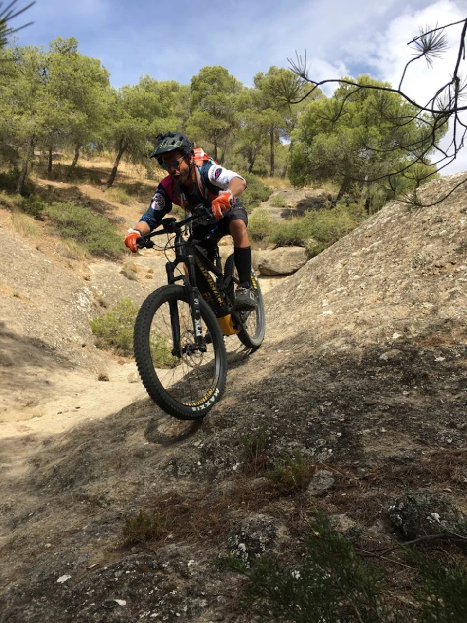 A cyclist wears protective gear, riding a mountain bike on a rocky trail surrounded by trees.