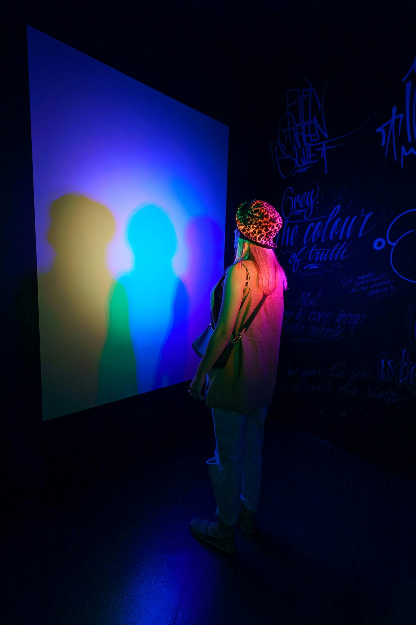 Eine Person mit einem Hut mit Leopardenmuster steht vor einer Wand, die bunte Schatten in einen dunklen Raum mit Graffiti wirft.