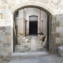 Stone archway leading to an enclosed courtyard with stone stairs, potted plants, and a dark doorway in the background.
