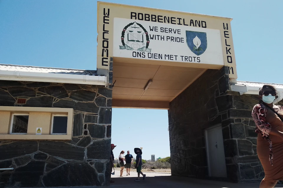 An entrance gate with "Robbeneiland" and "Welcome" displayed, people walking through, and one person wearing a mask on the right.