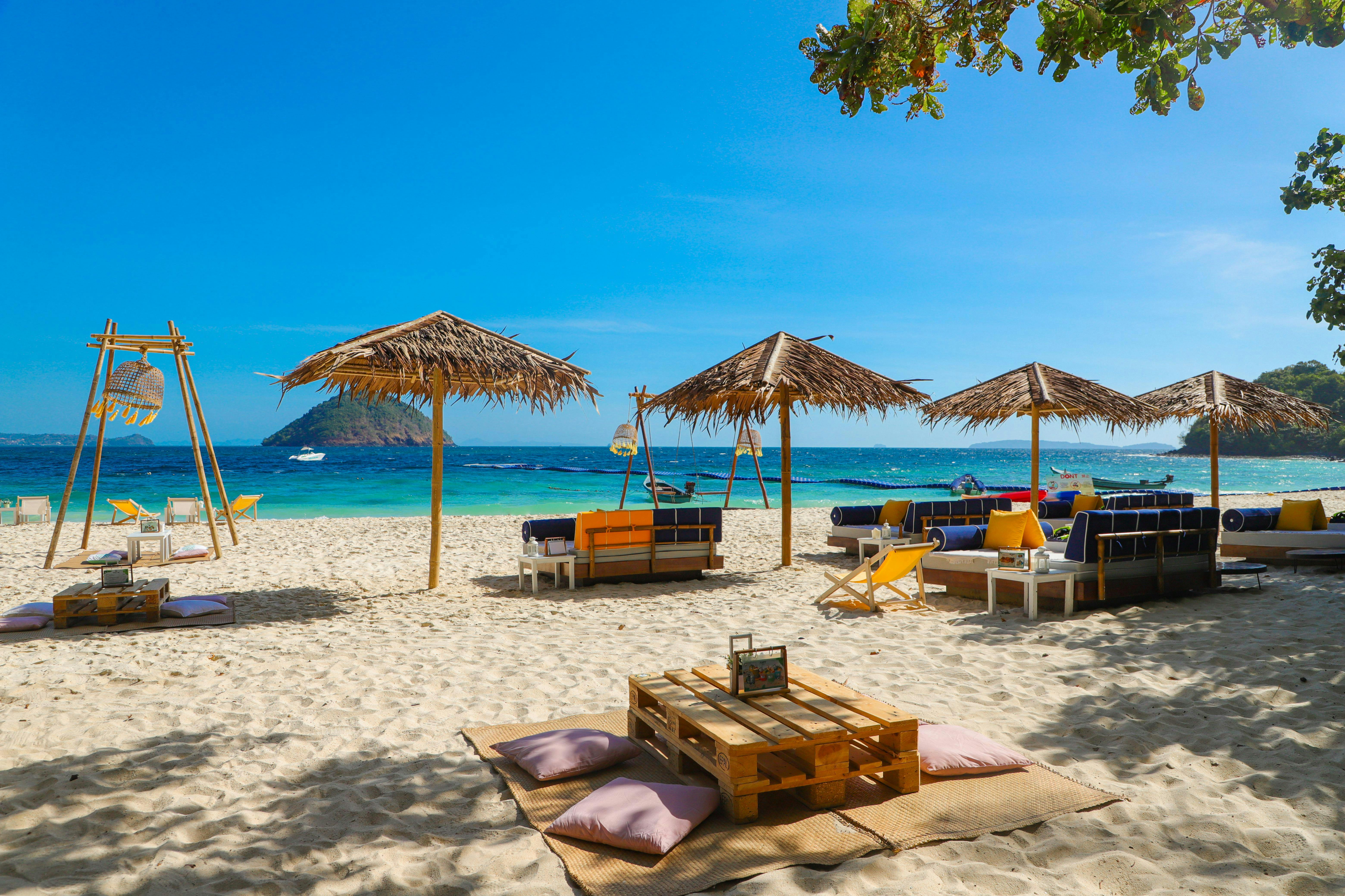 Пляжная сцена с соломенными зонтиками, шезлонгами, столами и качелями. Чистое голубое небо и океан с небольшим островом на расстоянии.