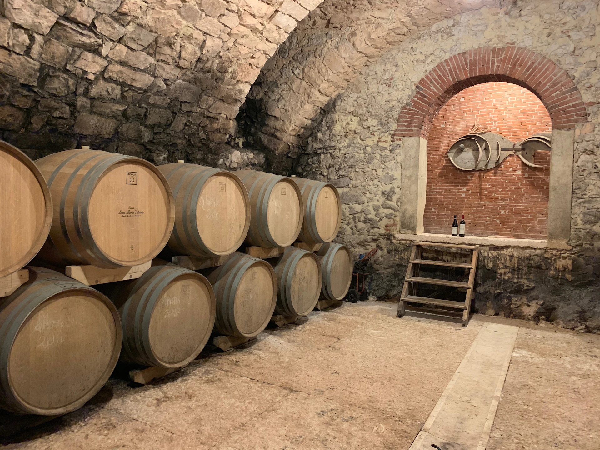 Stone cellar with stacked wooden barrels, a small staircase leading to a brick archway, and two wine bottles on a table.