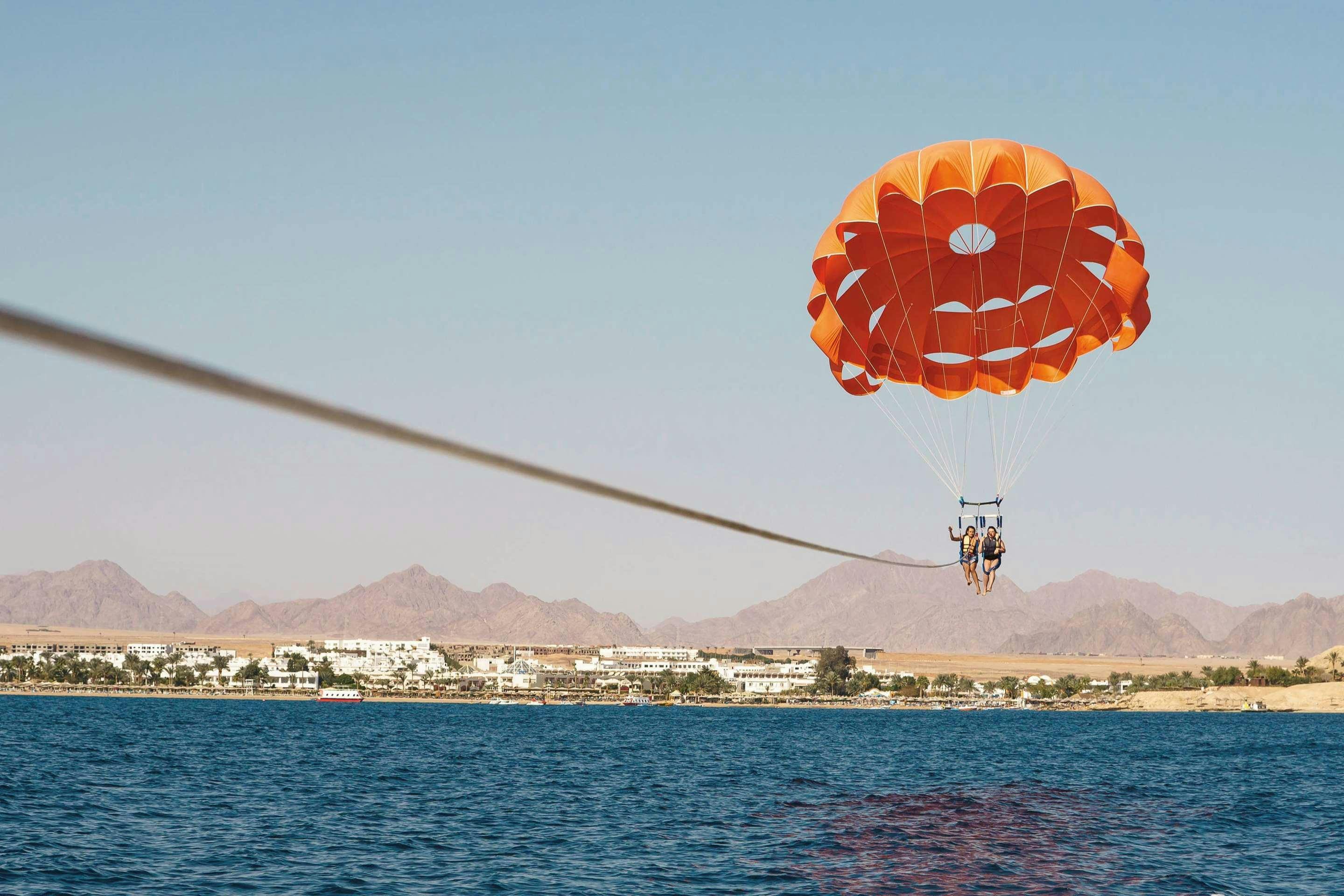 Two people parasailing with an orange parachute over blue water, with a coastal town and mountains in the background.