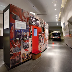 Museiutställning med politiska affischer, en lyssningsstation med hörlurar och en svart bil med en öppen bagagelucka i bakgrunden.