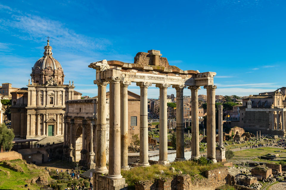 Ancient ruins with tall stone columns under a blue sky, surrounded by historic buildings and greenery.
