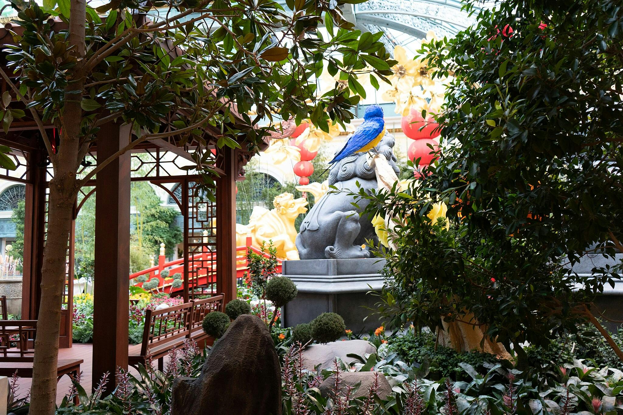 Svěží zahrada s červeným dřevěným altánem, kamennou sochou zvířete a modrým ptákem na vrcholu sochy, obklopená různými rostlinami a stromy.