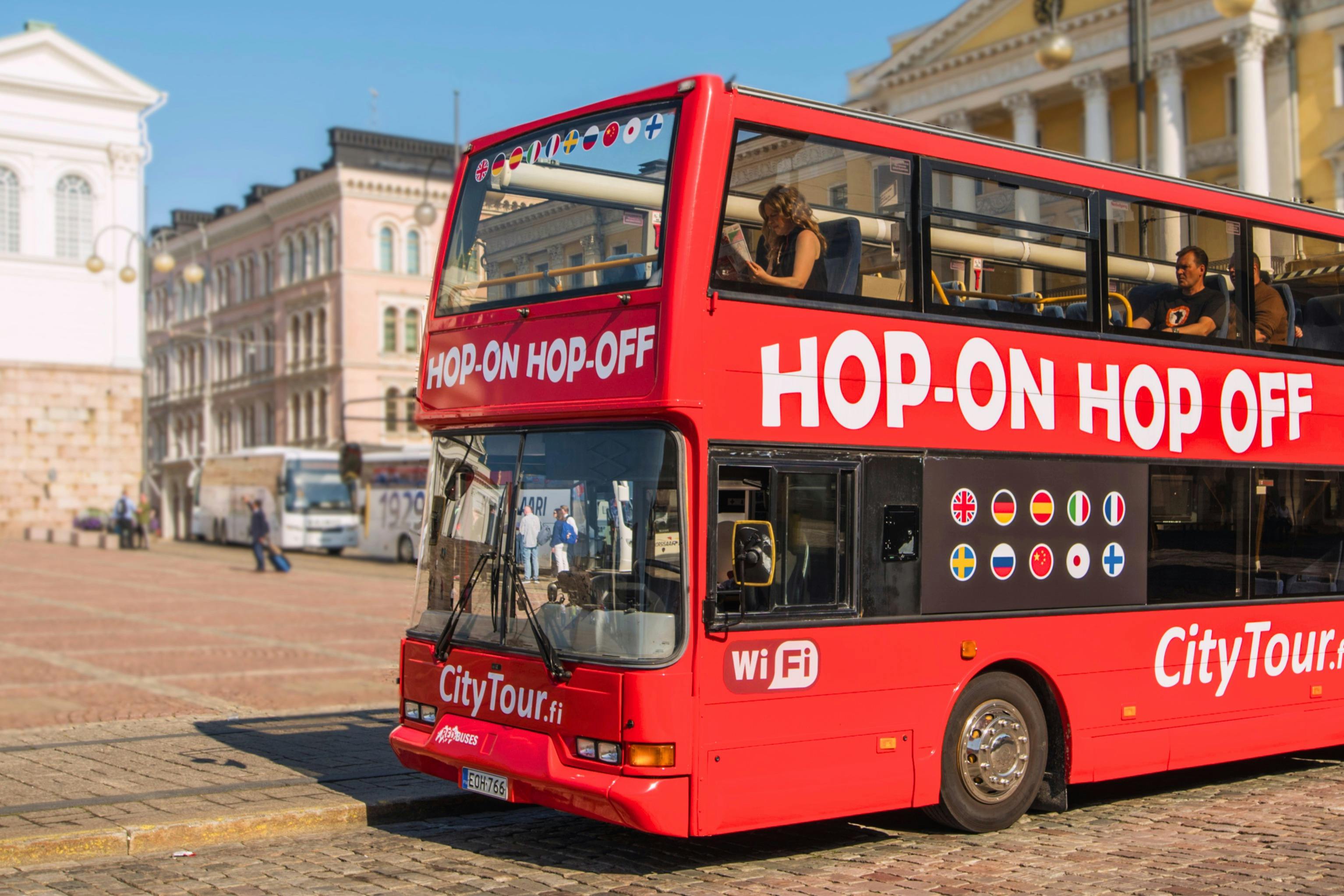 「乗り降り自由」と書かれた街の広場にある真っ赤なダブルデッカーツアーバス。賑やかな街の背景。