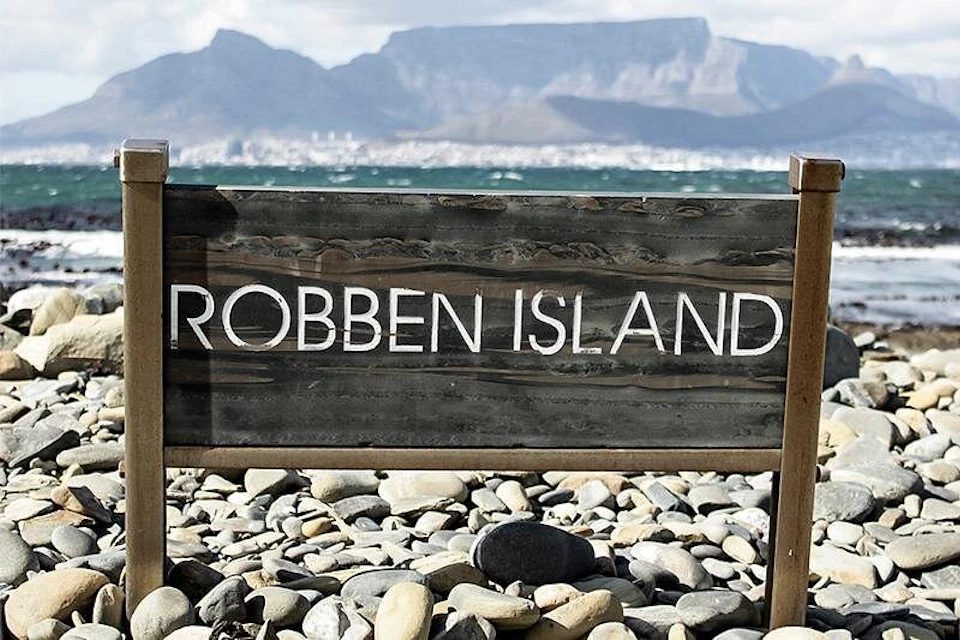 A wooden sign reading "Robben Island" stands on a rocky shoreline with mountains and a body of water in the background.
