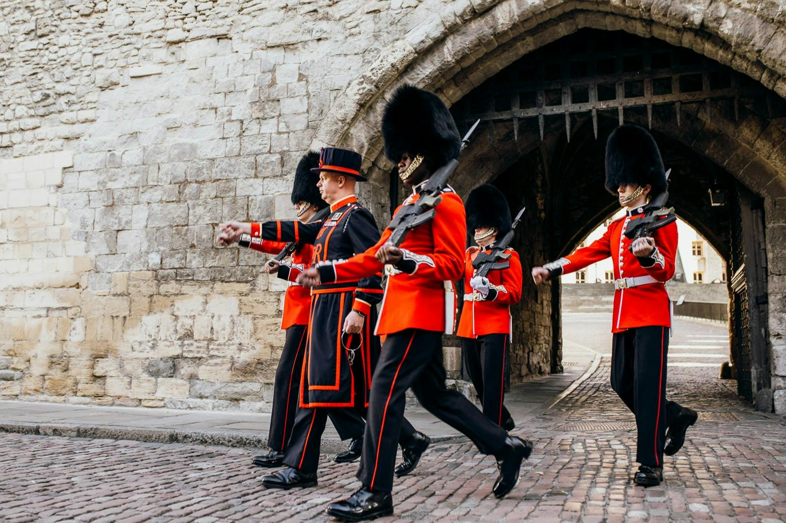 Guards in red uniforms and tall black hats march in formation near a stone archway.