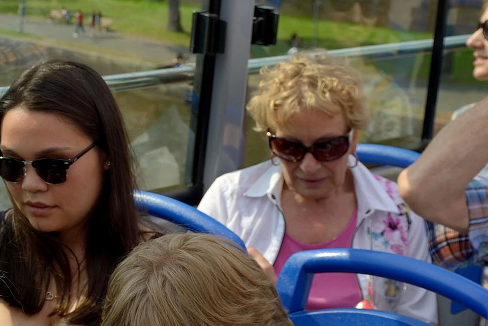 A group of people, including a young boy and women taking a selfie, ride on an open-top tour bus with a cityscape in the background.