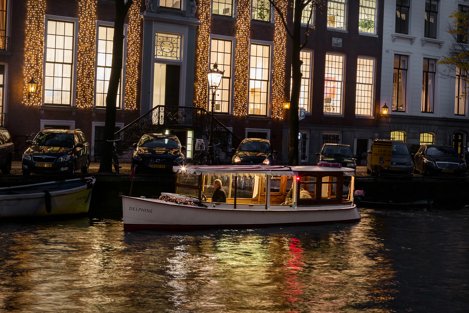 A boat named "Delphine" on a canal with lit buildings behind it and parked cars along the water's edge at dusk.