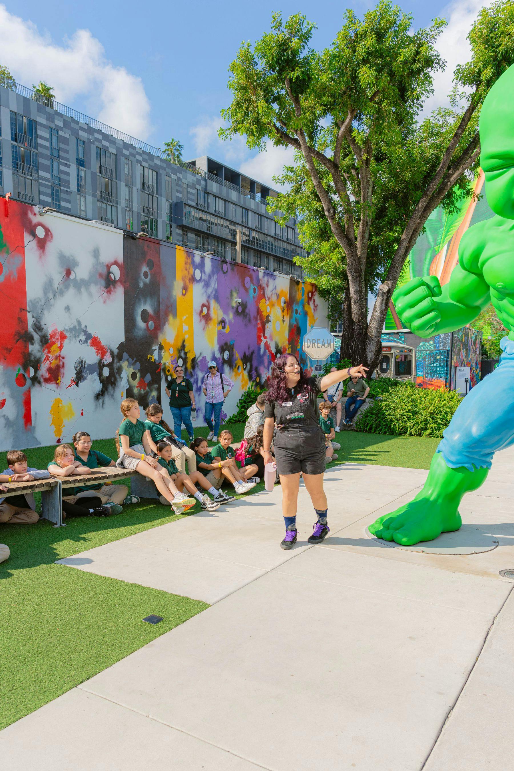一群孩子全神贯注地坐在人造草坪上，一名妇女指着城市公园里一座绿色的大型绿巨人雕像。