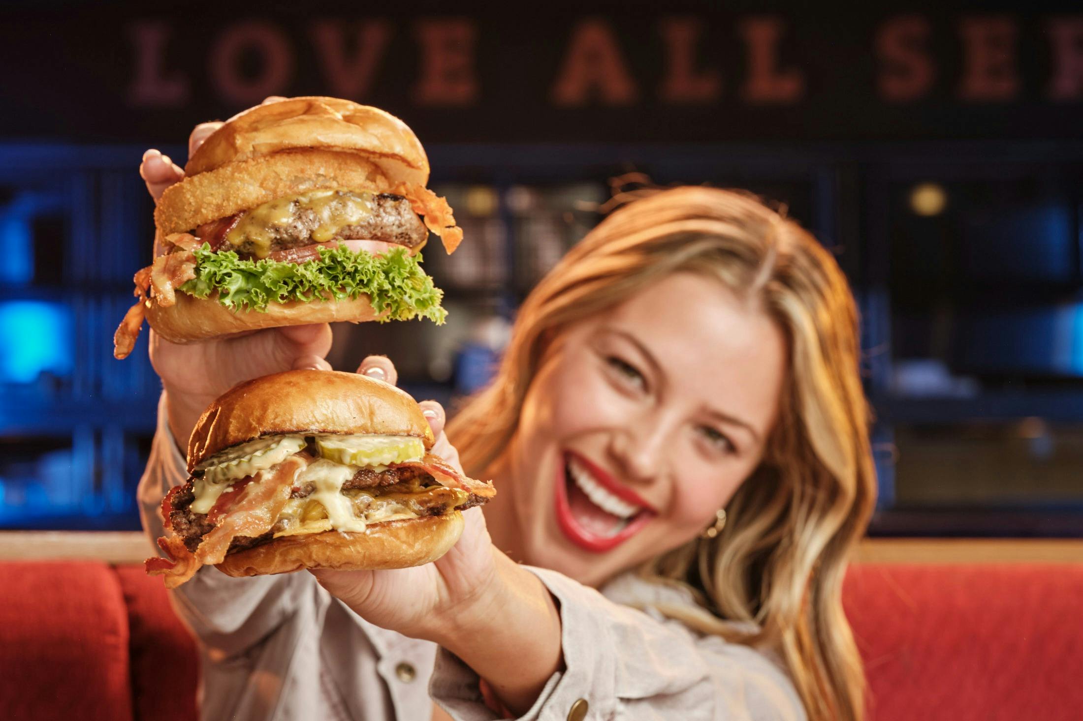 Una persona somrient amb dues hamburgueses, una a cada mà, amb el fons d'un restaurant.