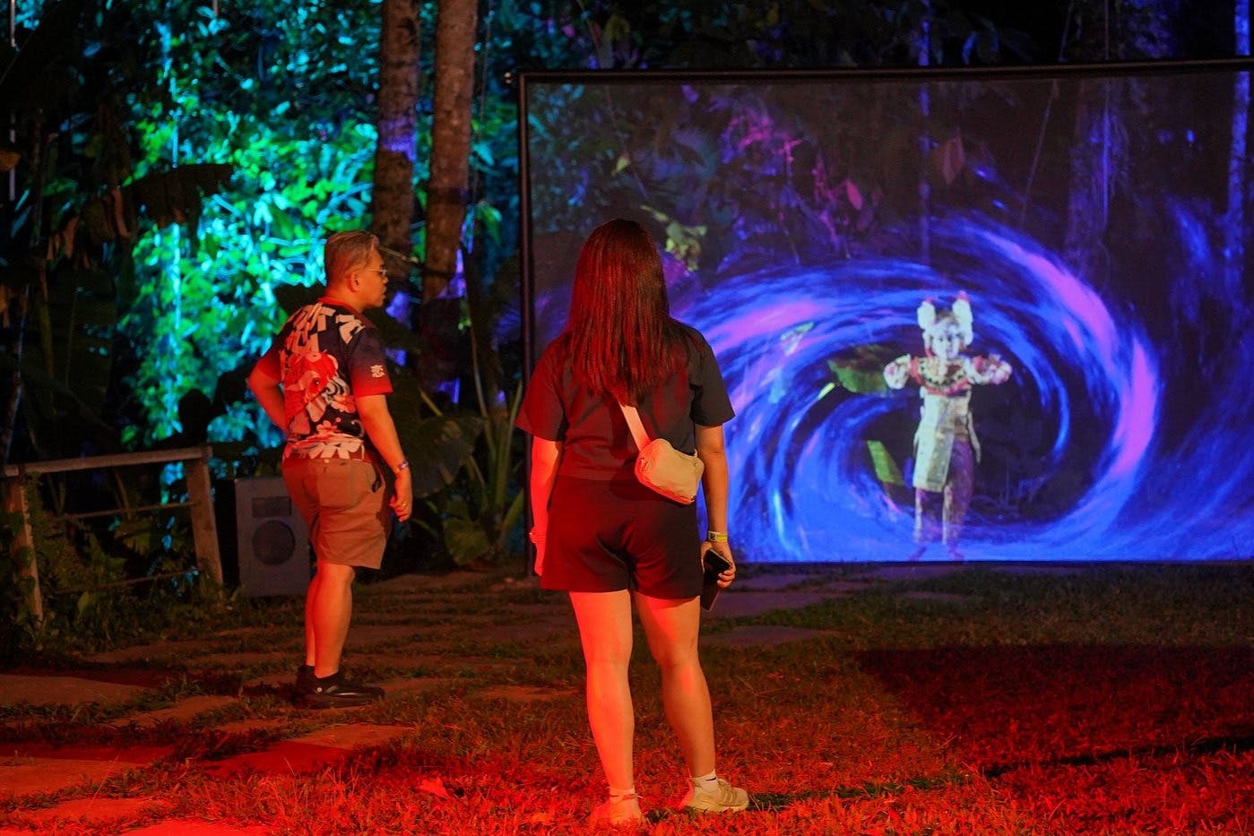 2人の人物が大きなスクリーンの前に立っており、緑豊かな緑に囲まれた屋外環境で鮮やかなデジタル画像を表示しています。