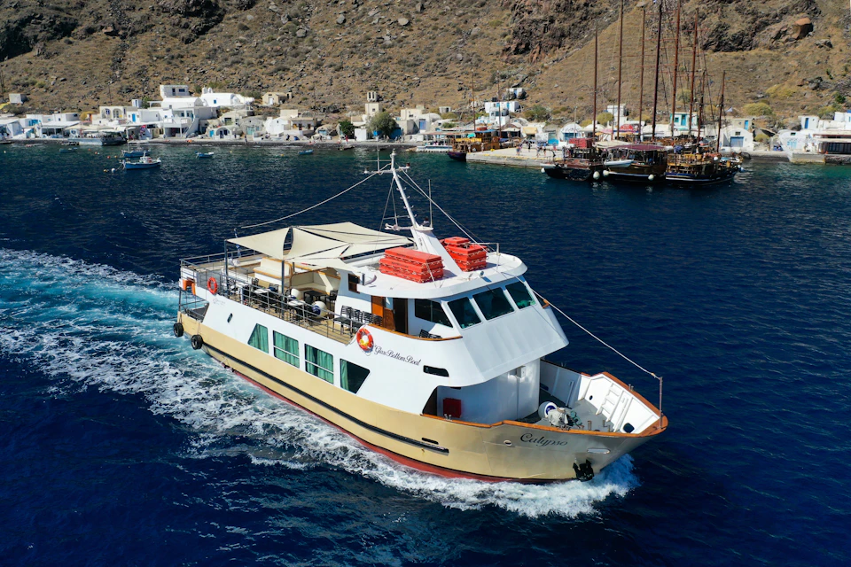 A boat named Calypso sails in blue waters near a coastal town with white buildings and boats docked by the shore.
