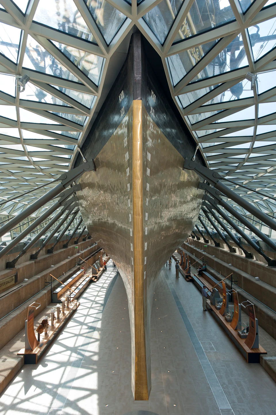 A large ship's hull displayed indoors within a glass-roofed structure, flanked by exhibits and railings.