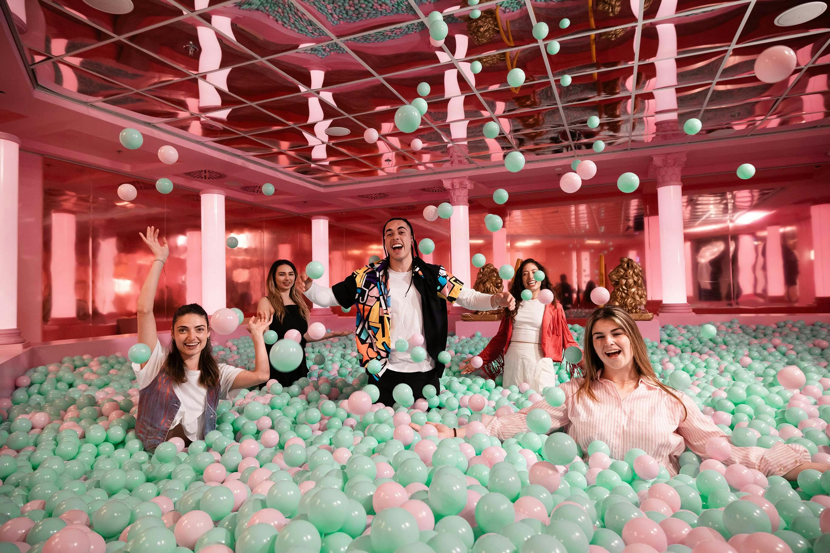五个人在一个充满粉红色和绿色球的球坑里微笑着玩耍，周围是粉红色的墙壁和镜面天花板。