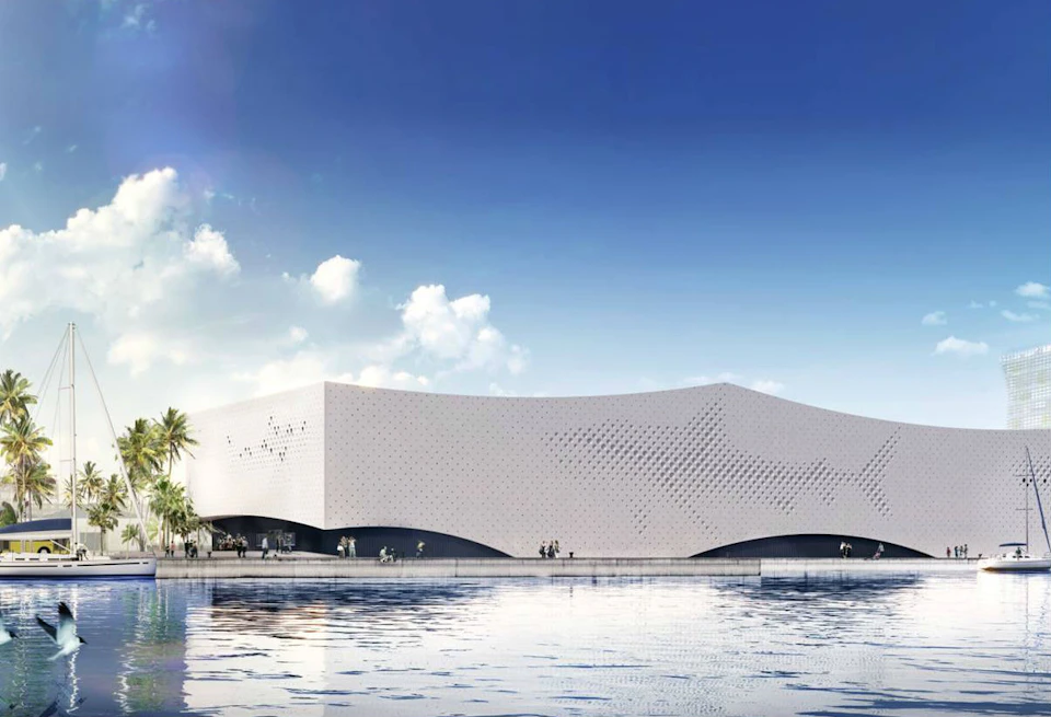 Modern white building with perforated design, reflecting in a calm water surface, under a blue sky with scattered clouds.