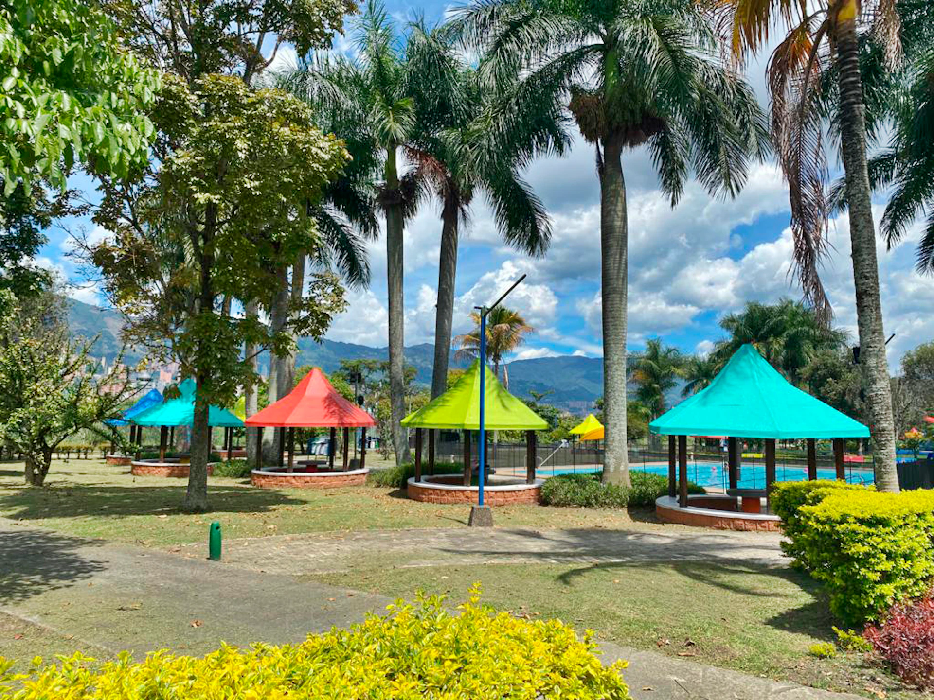 Colorful gazebos under tall palm trees in a park with mountains in the background.